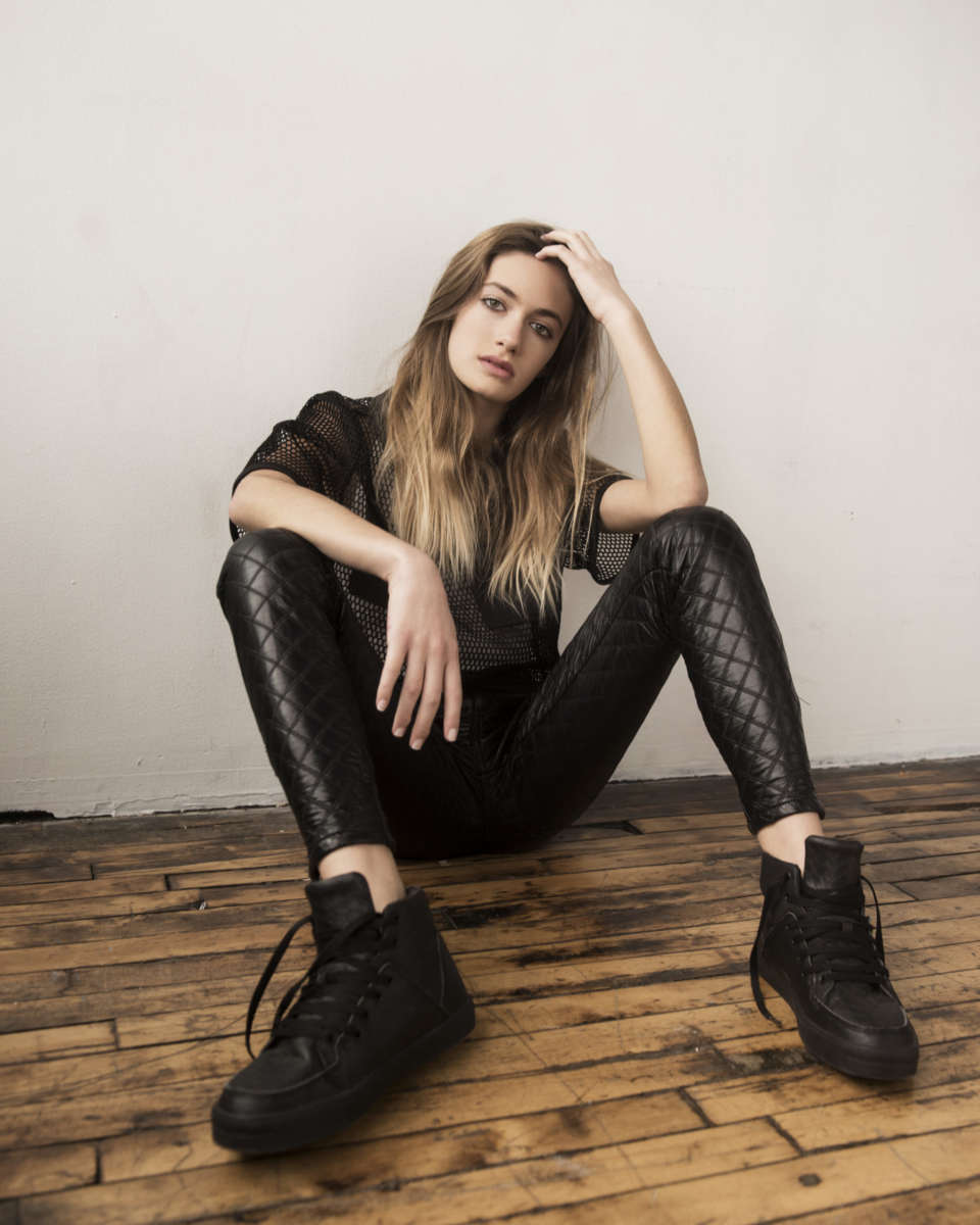 Actress and Singer Nicole Berger, seated on the ground, full body shot, wearing black leather pants, a black mesh top and black sneakers. On hand in her hair the other on her propped up knee. Shot by Matthew Priestly