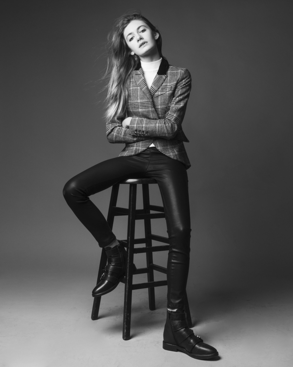 Actress and Singer Nicole Berger, black and white full body shot wearing a plaid blazer, black leather pants and black boots. Arms crossed and chin tilted slightly upwards. Hair, straight, pulled to the left over the shoulder. Sitting on a stool, one foot slightly lifted. Shot by Matthew Priestly