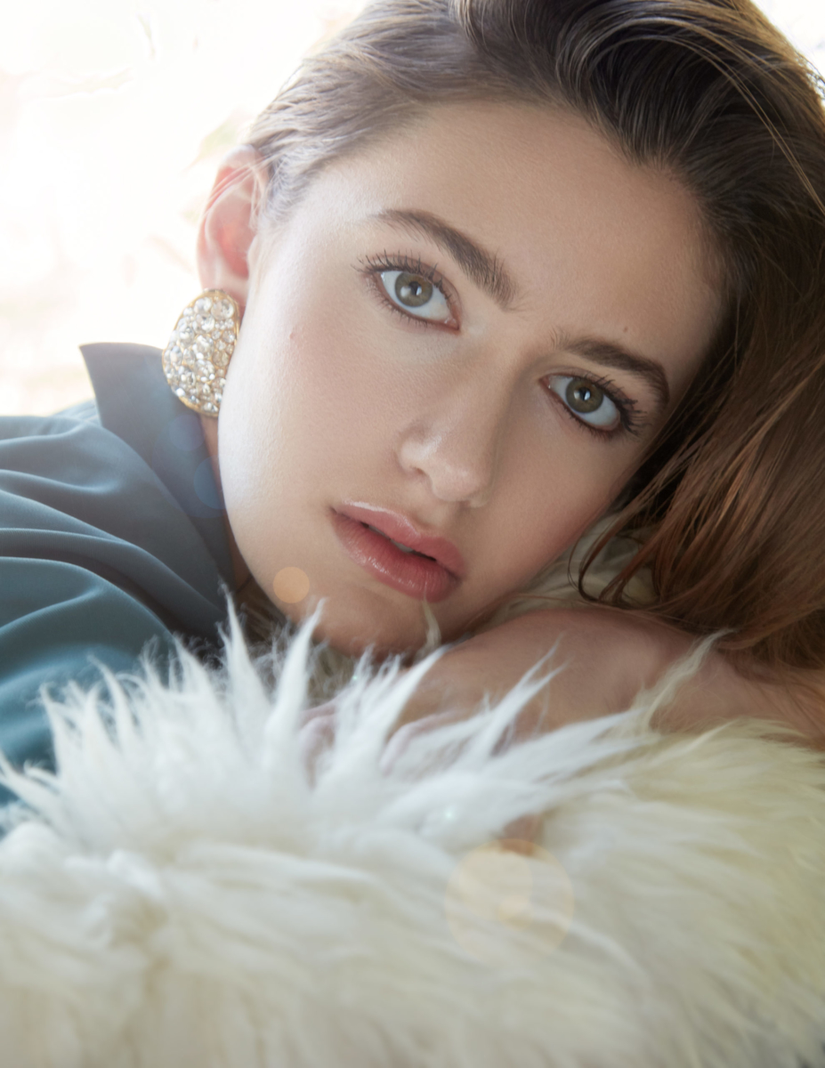 Actress and Singer Nicole Berger, close up headshot with her hair pulled over her head, wearing a turquoise blue blouse and chunky earing. Pink lip. Laying on a white faux fur pillow. Shot by Beau Nelson.