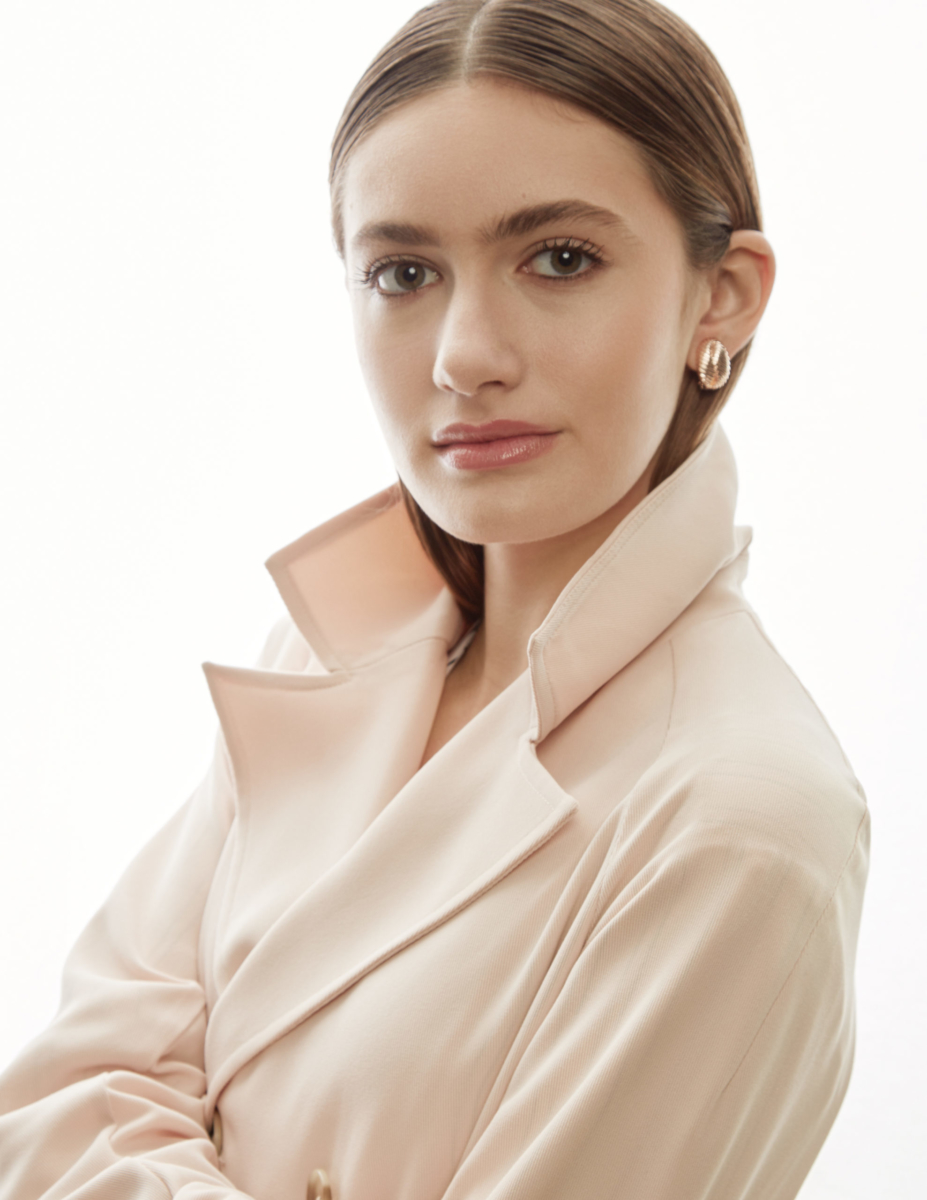 Actress and Singer Nicole Berger, wearing a beige silk jacket with the collar popped. Her hair is slicked back and parted down the middle. Wearing gold earrings. Body in profile but she is looking straight into the camera. Makeup is natural. Shot by Beau Nelson